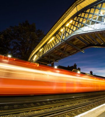 Train Passing Under a Bridge at Night A red train moving under a lit bridge at a railway station during nighttime.
